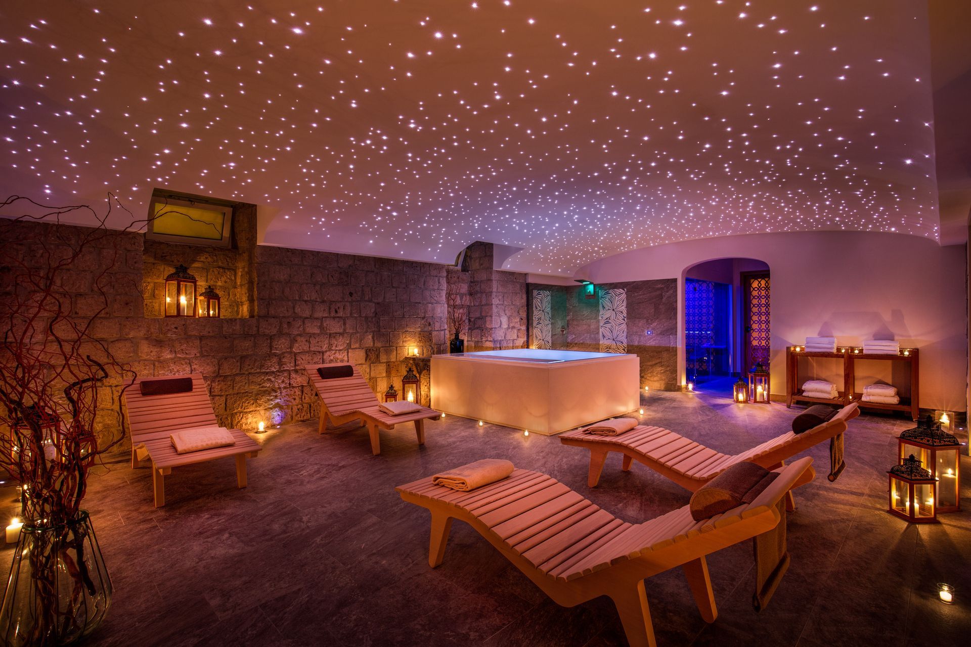 A dimly lit, stone-walled spa room with a star-light ceiling, a central hot tub, and multiple lounge chairs.
