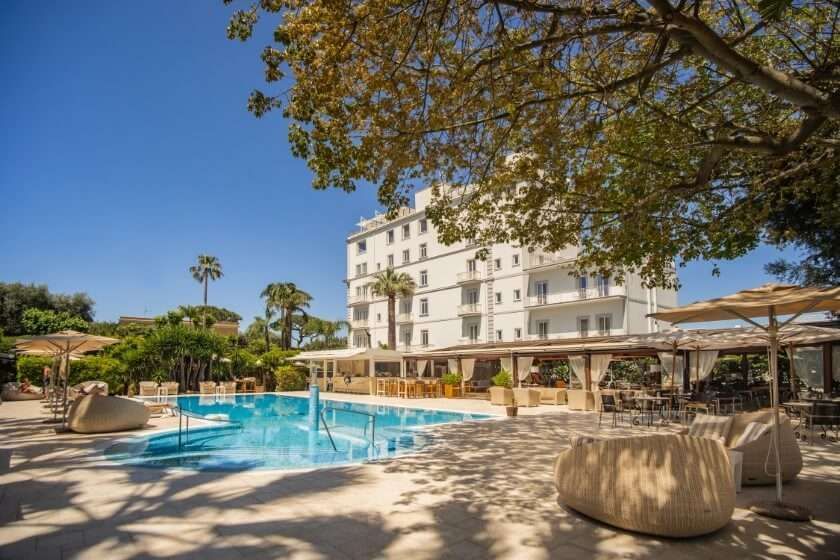 Una piscina all'aperto in un luminoso hotel a più piani, circondato da alberi e lettini all'ombra, in una giornata di sole.
