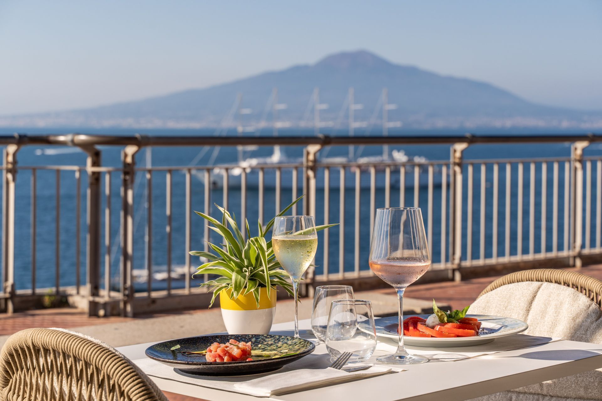 Tavolo soleggiato in riva al mare con bicchieri da vino, frutta e pianta in vaso, con vista sulla baia