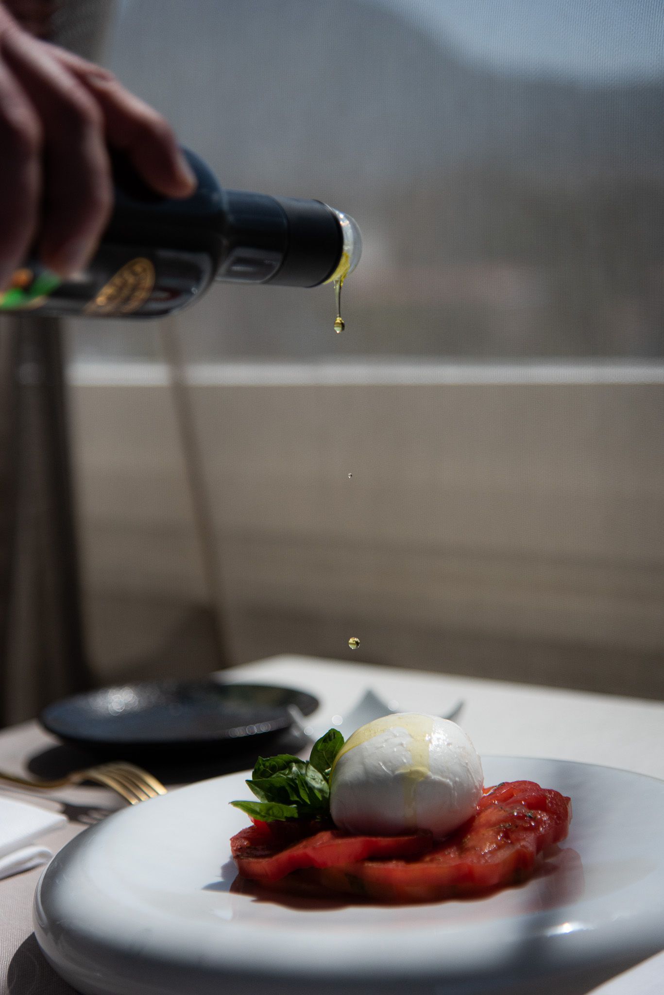 A hand pours olive oil from a dark bottle onto a fresh mozzarella and tomato salad on a white plate.