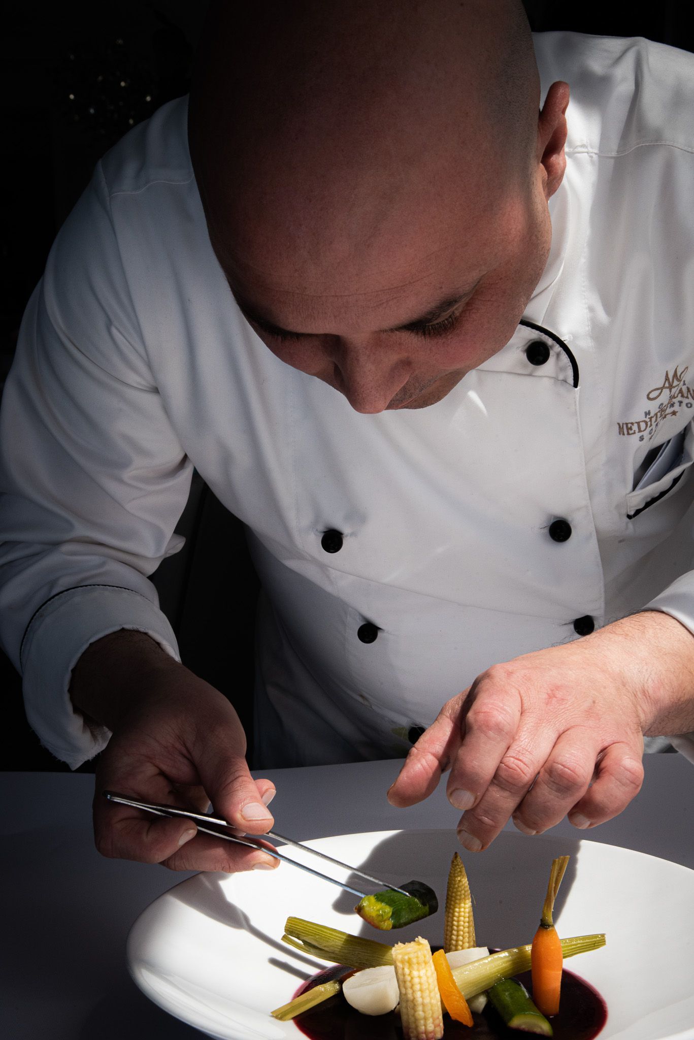 A chef in a white uniform uses tweezers to carefully arrange small vegetables on a plate in a dark setting.