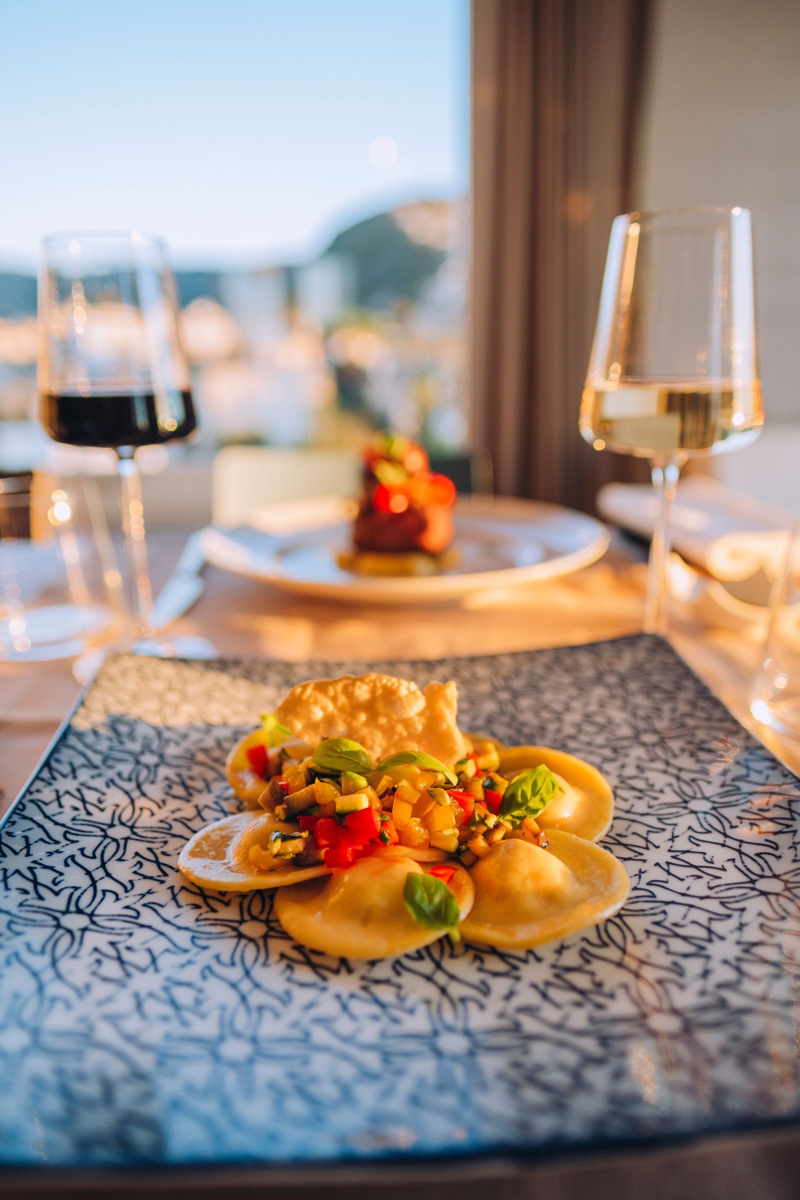 A dish of ravioli topped with vegetables on a decorative plate, set on a table with wine glasses and a blurred background.