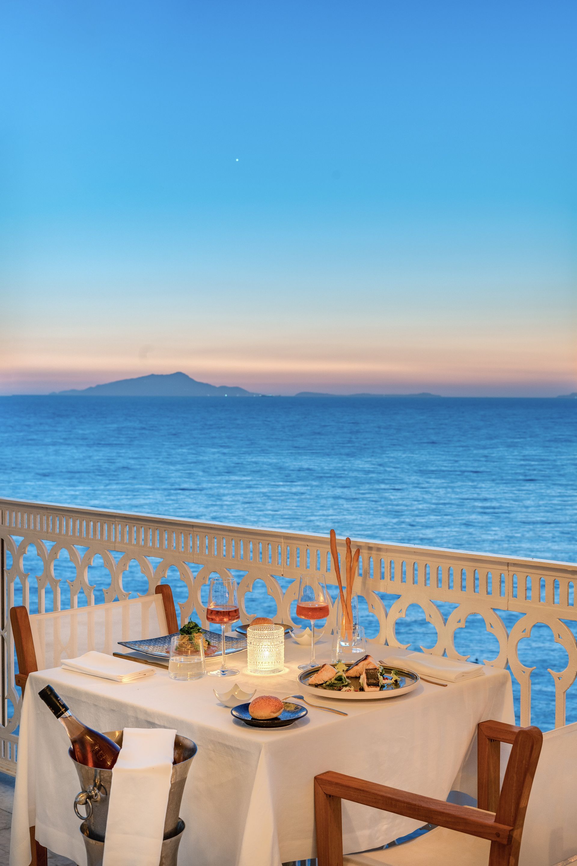 A romantic table set for two on a balcony overlooking the ocean at sunset with a bottle of wine in a cooling bucket.