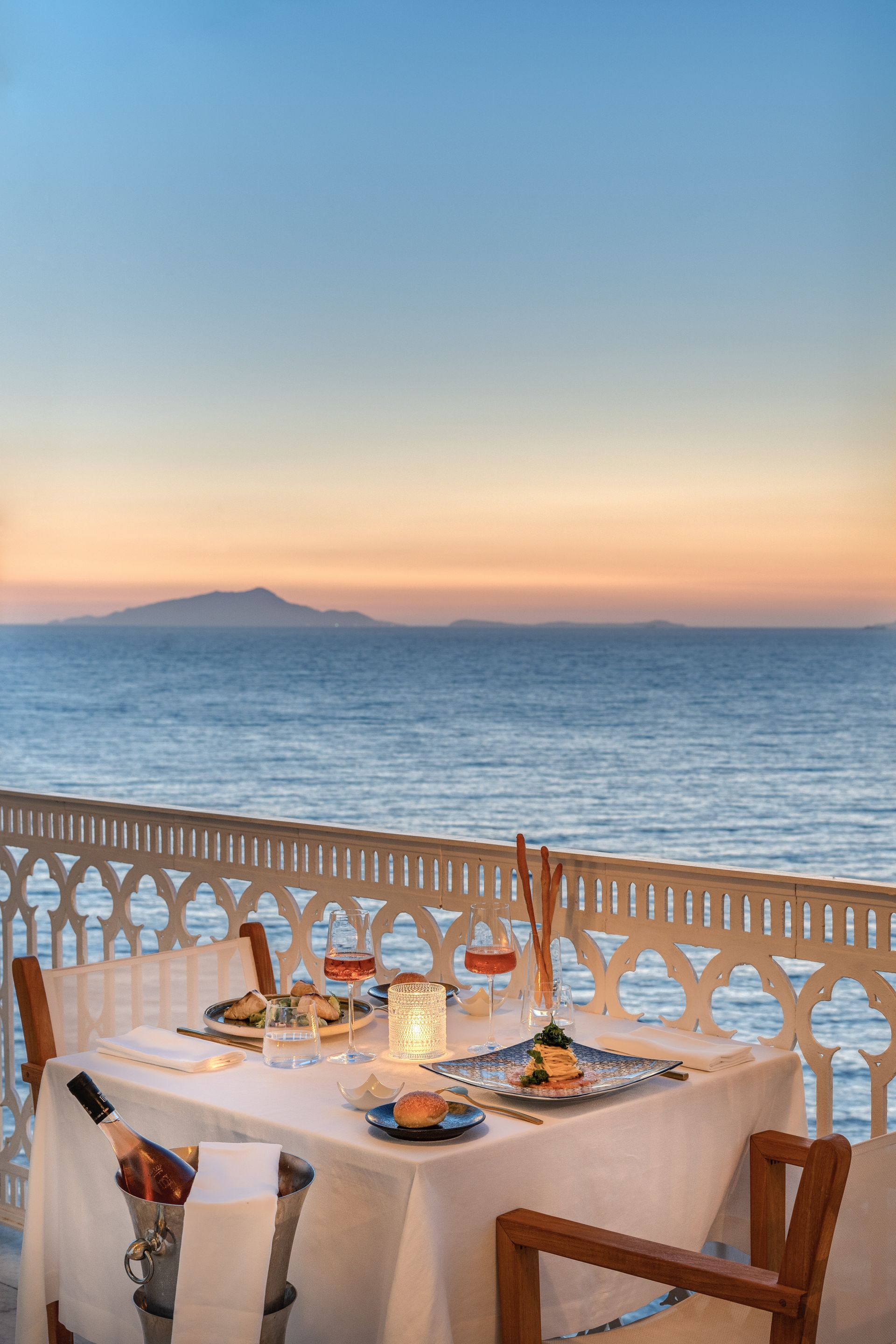 A table set for dinner on a balcony overlooking the sea at sunset, with a bottle of wine in a cooler.