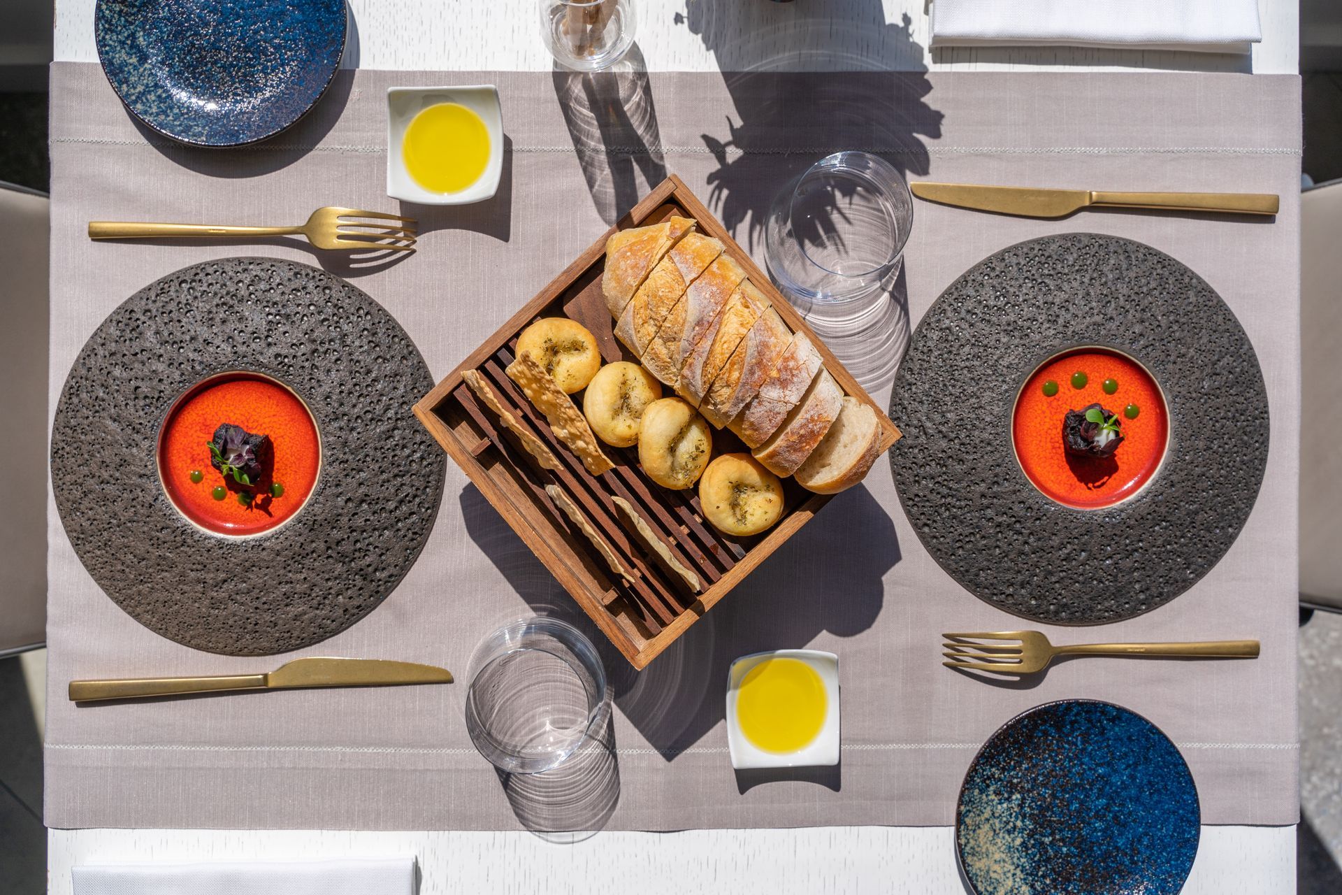 A table set for two with textured grey plates holding red soup, a central basket of bread, gold cutlery, and olive oil.