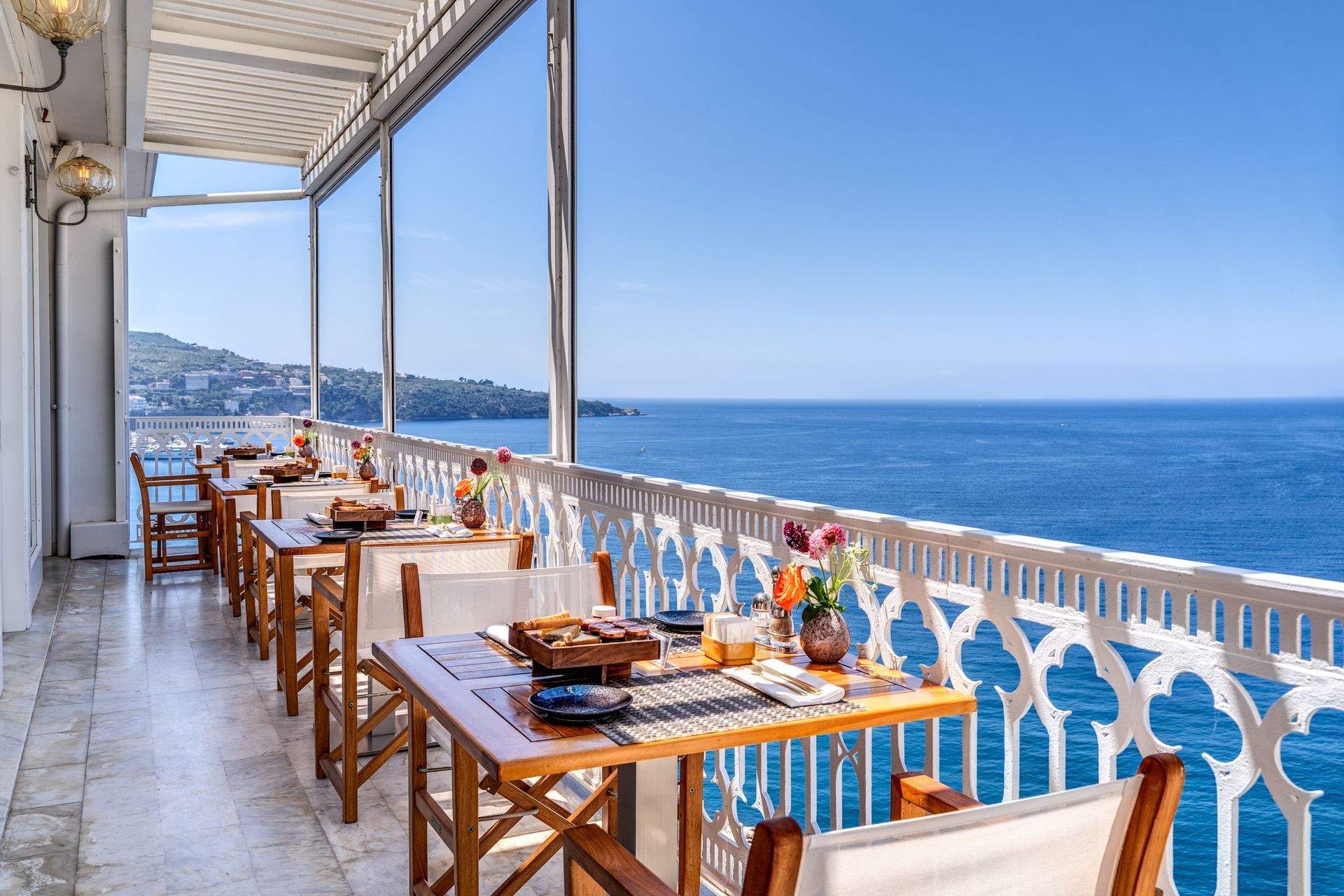Un ristorante con balcone soleggiato, tavoli in legno e sedie bianche, affacciato su un mare calmo e di un blu intenso.