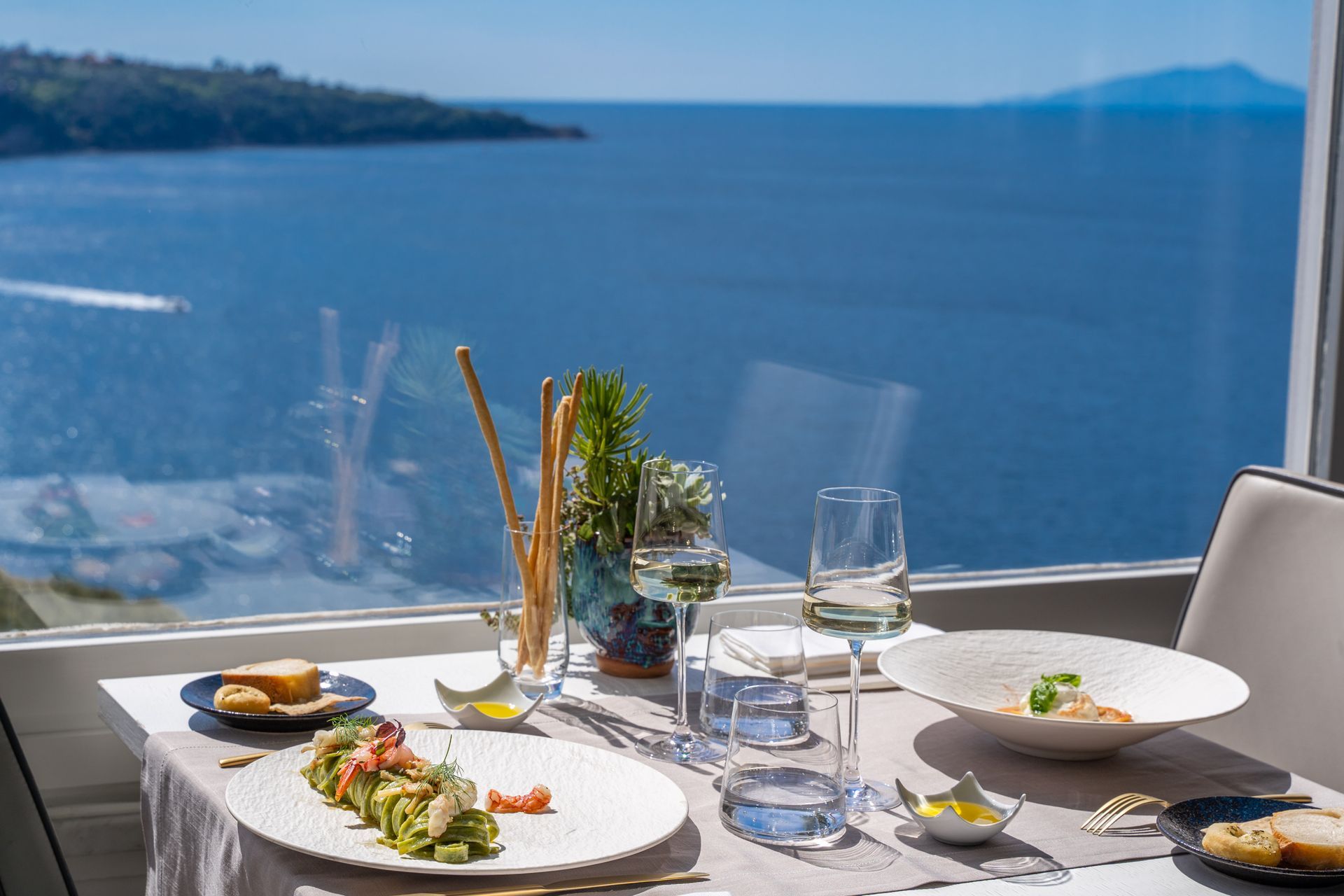 A table set for a meal with pasta, breadsticks, and wine, overlooking a blue ocean and distant island on a sunny day.