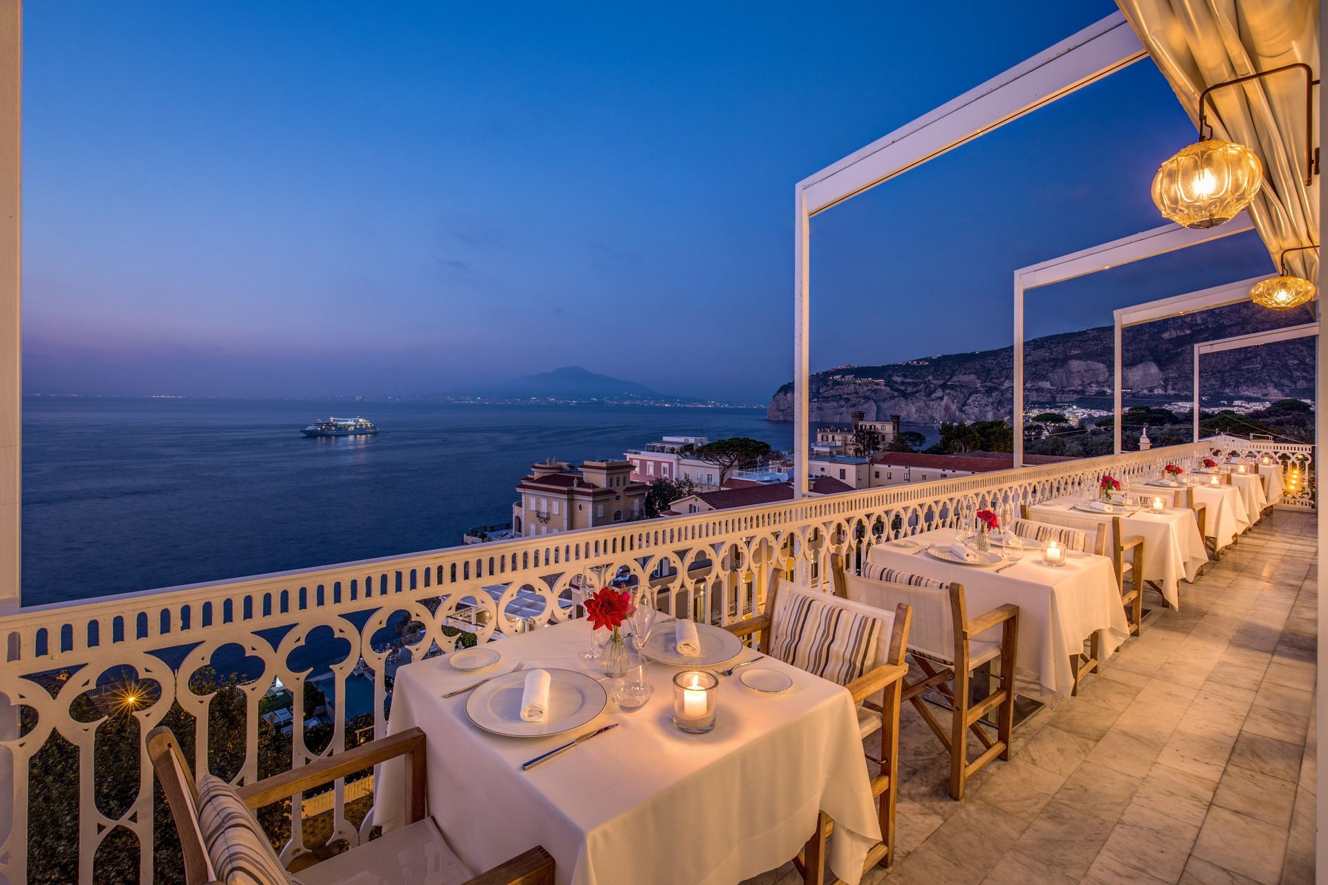 La terrazza di un ristorante al tramonto, affacciata su un mare calmo, con tavoli e sedie apparecchiati con tovaglie bianche e lampade a sospensione a forma di globo.