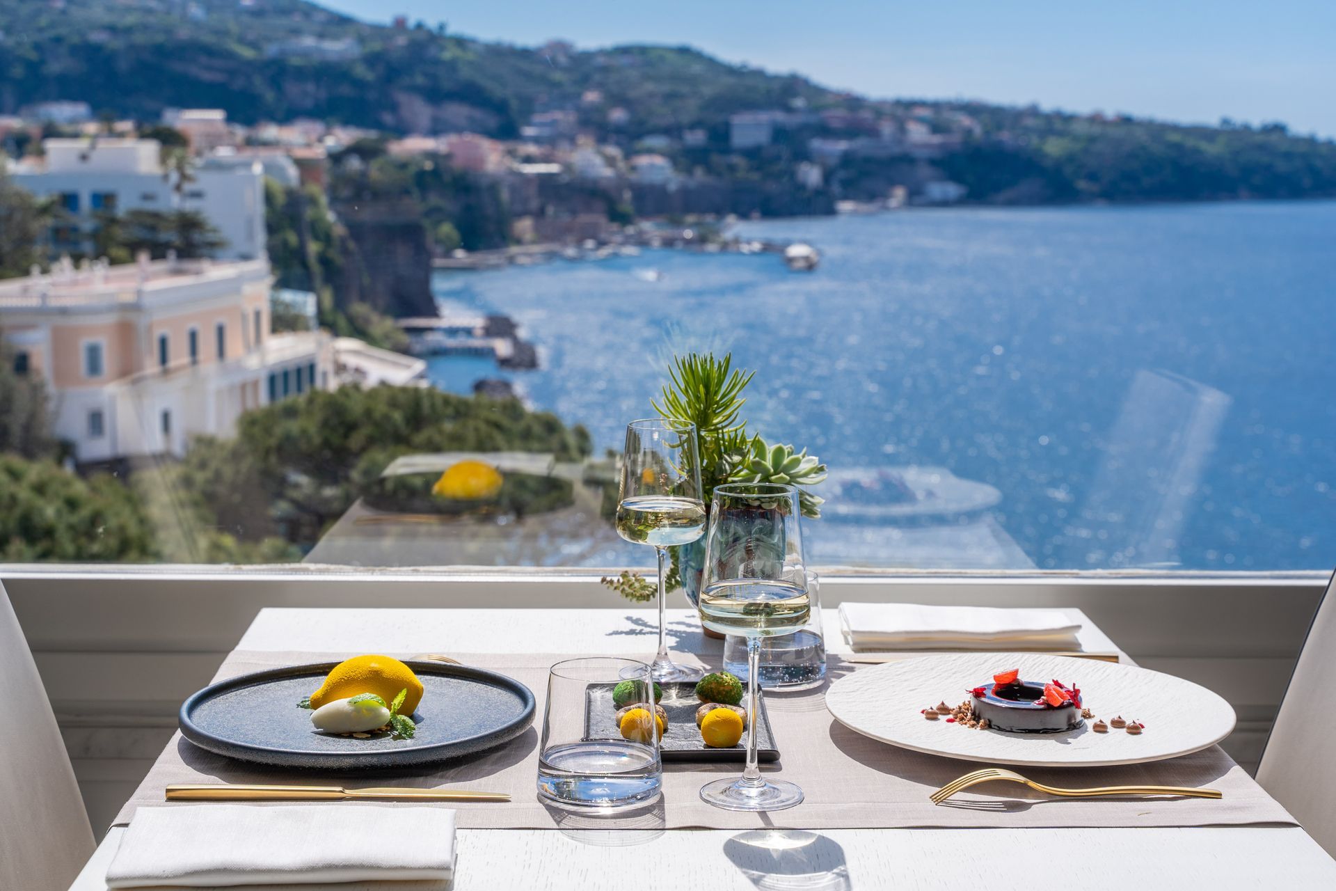 Tables set for dinner on a balcony overlooking the ocean at twilight, with white tablecloths and warm glowing lights.