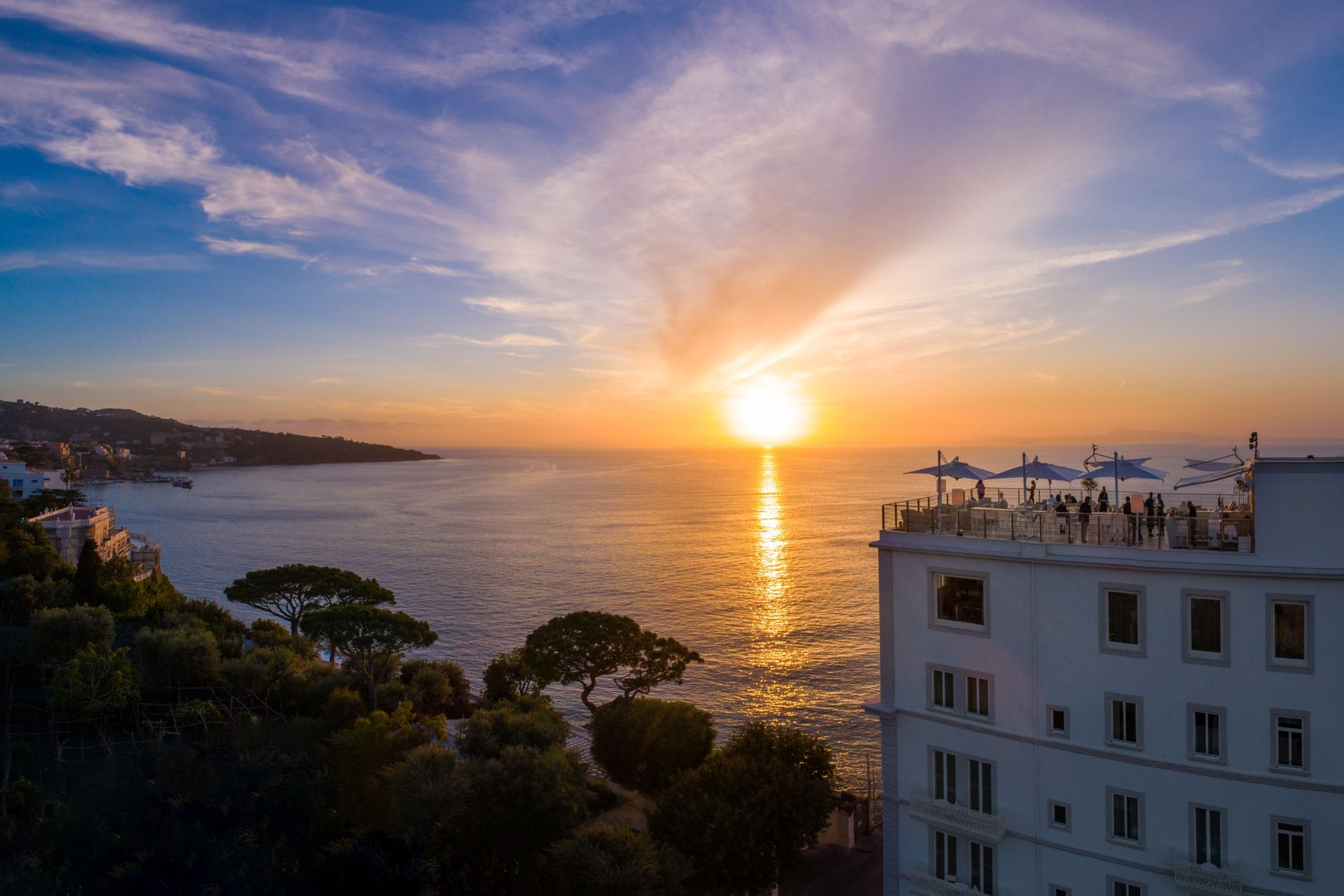 Un hotel bianco affacciato su un oceano calmo durante un tramonto dorato, con persone sedute su una terrazza panoramica.