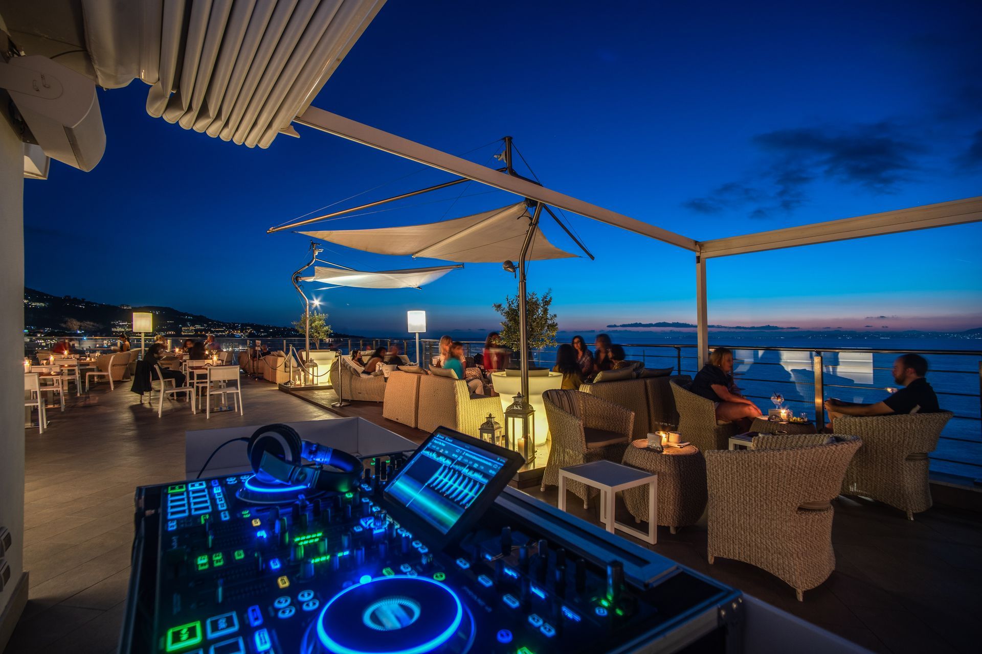 In primo piano, attrezzature da DJ che si affacciano su una terrazza panoramica all'aperto al crepuscolo, con persone sedute in riva all'oceano.