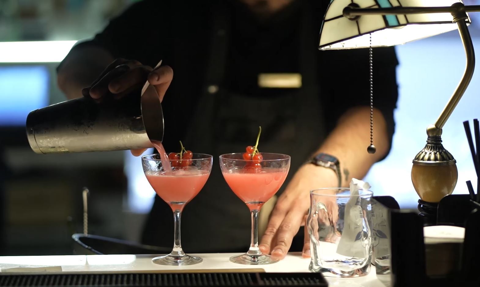 A bartender pours a pink cocktail into a stemmed glass next to another already garnished with a small red pepper.