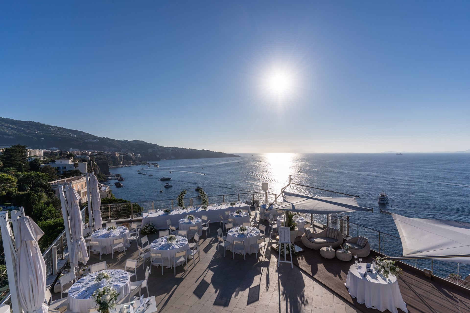 Terrazza esterna con vista sul mare al tramonto, arredata con tavoli apparecchiati con tovaglie bianche e ombrelloni per eventi.
