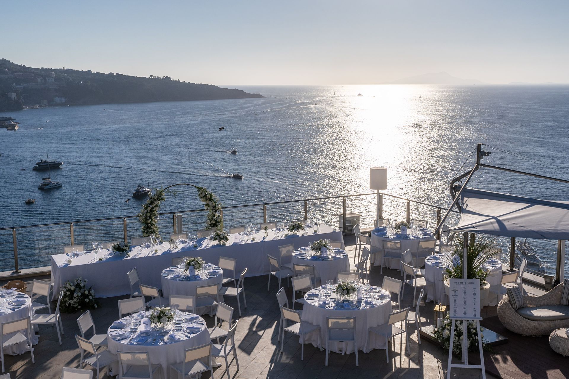 Una location per matrimoni all'aperto con tavoli apparecchiati con tovaglie bianche che si affacciano su un mare illuminato dal sole, con barche all'orizzonte.