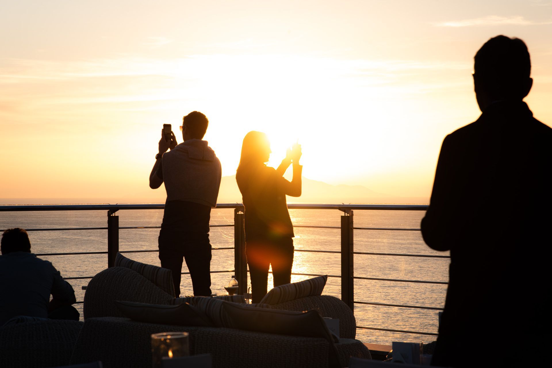 People watch a sunset from a deck overlooking the sea, with two individuals capturing photos with their smartphones.
