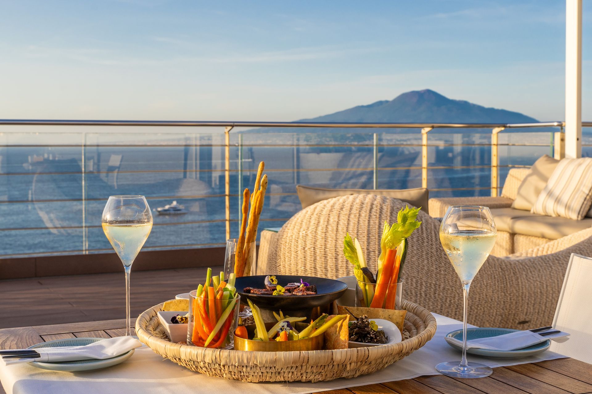 A food platter with appetizers and two glasses of wine on a balcony overlooking the sea and Mount Vesuvius at sunset.