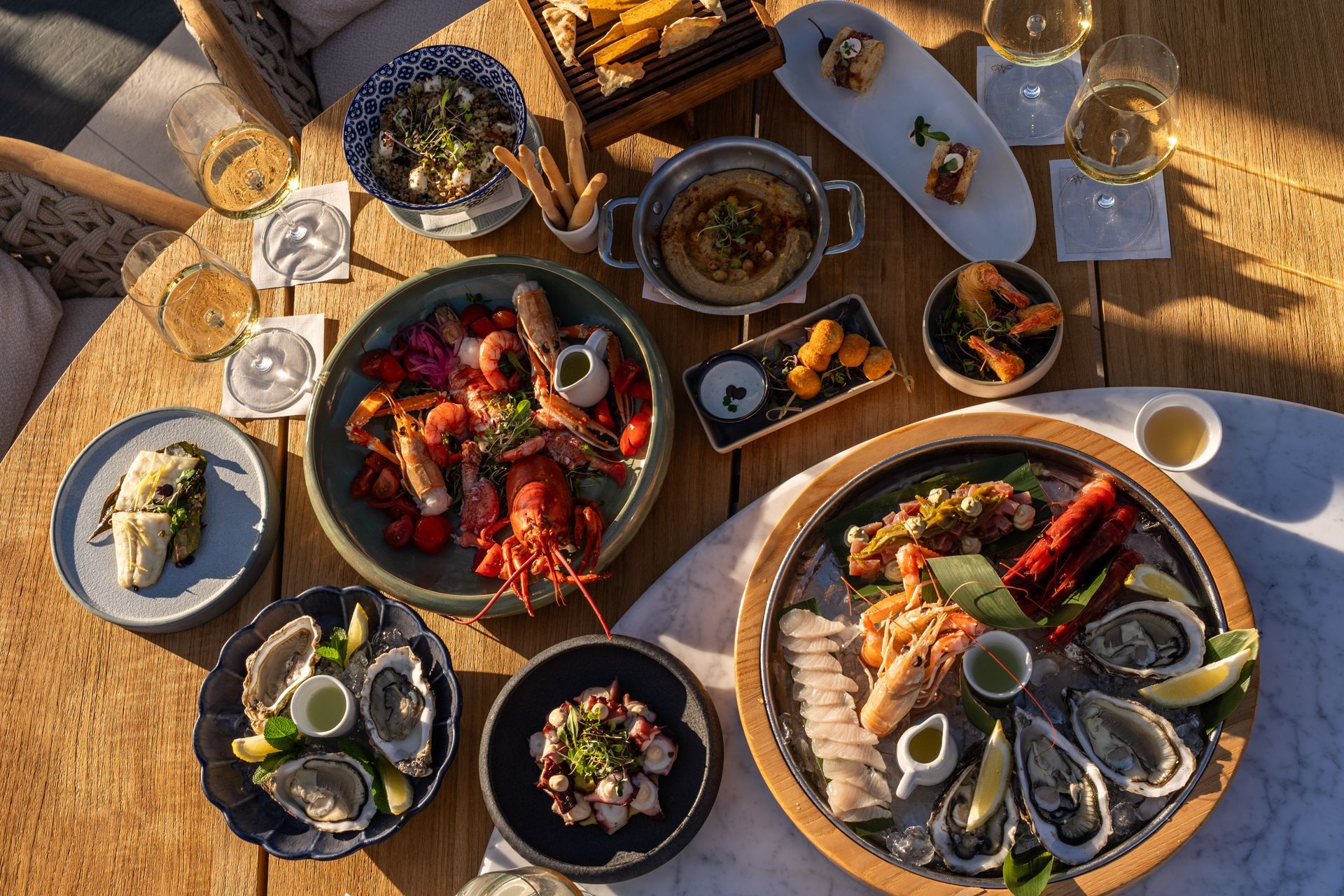 Un banchetto a base di frutti di mare imbandito su un tavolo illuminato dal sole, con aragoste, ostriche, gamberi e calici di vino.