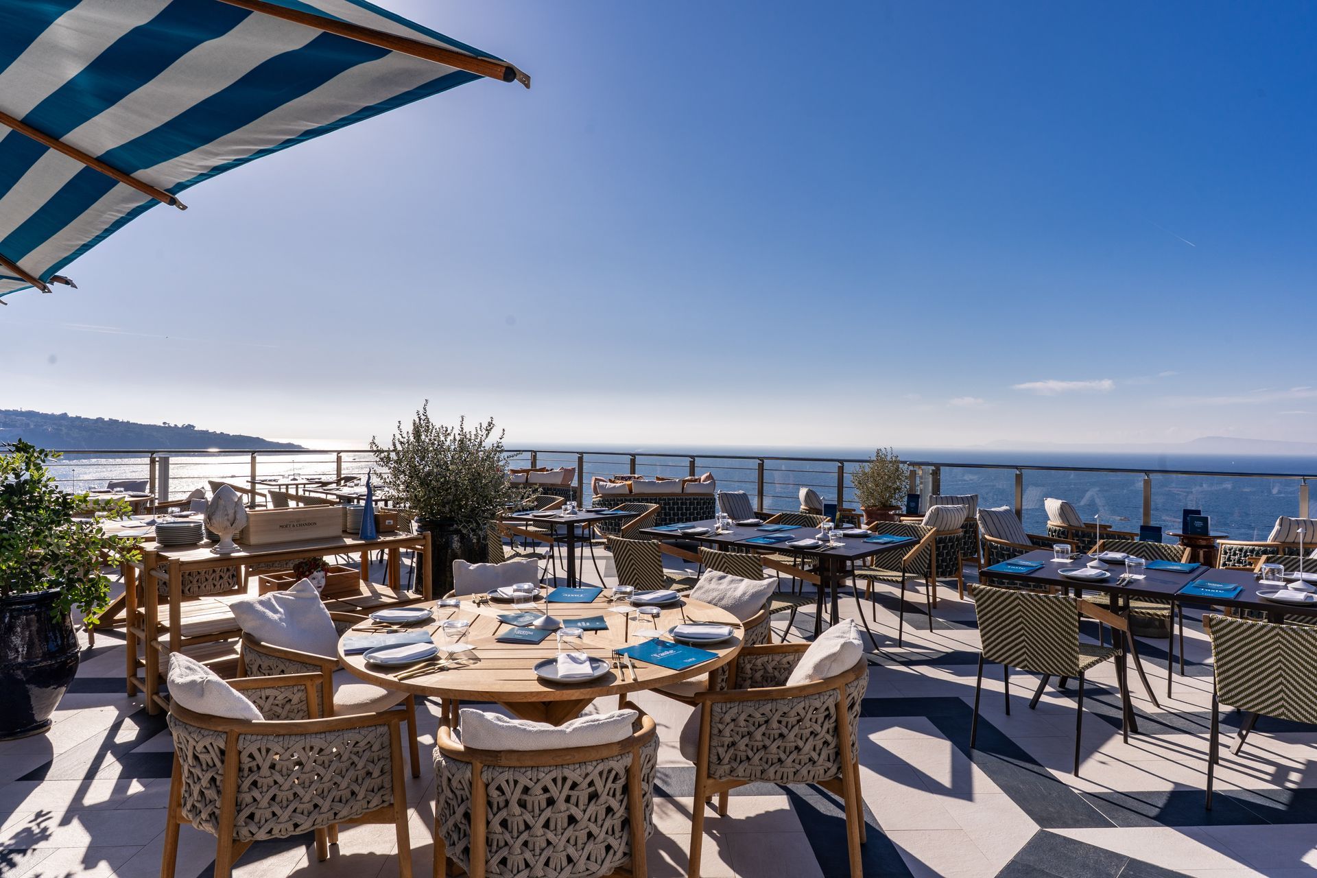 Outdoor lounge on a balcony overlooking the sea, with wicker furniture, white umbrellas, and white lanterns at sunset.