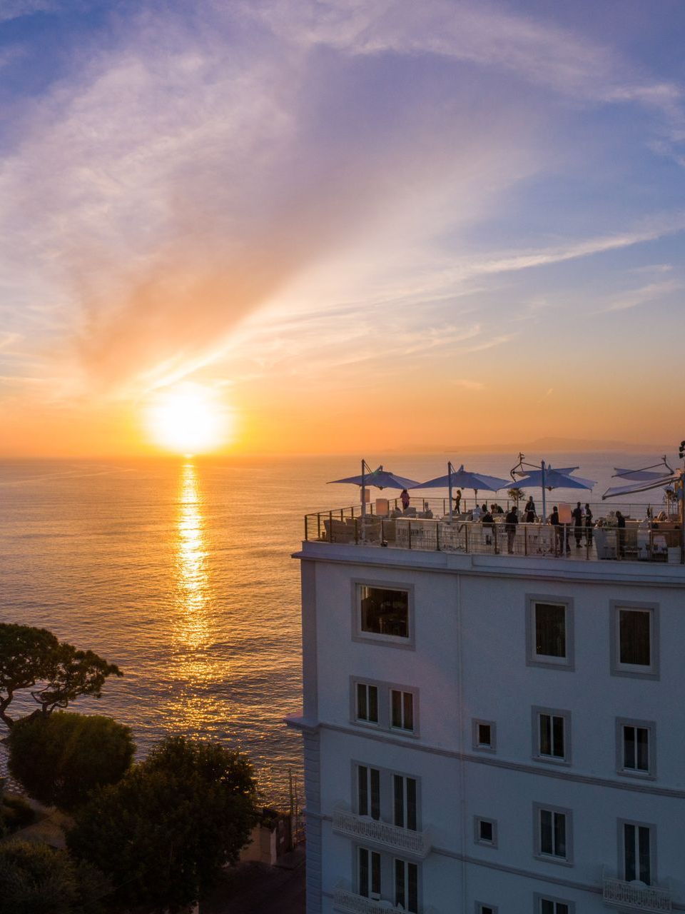 Un edificio alberghiero bianco con una terrazza sul tetto affacciata sull'oceano durante un tramonto dorato.