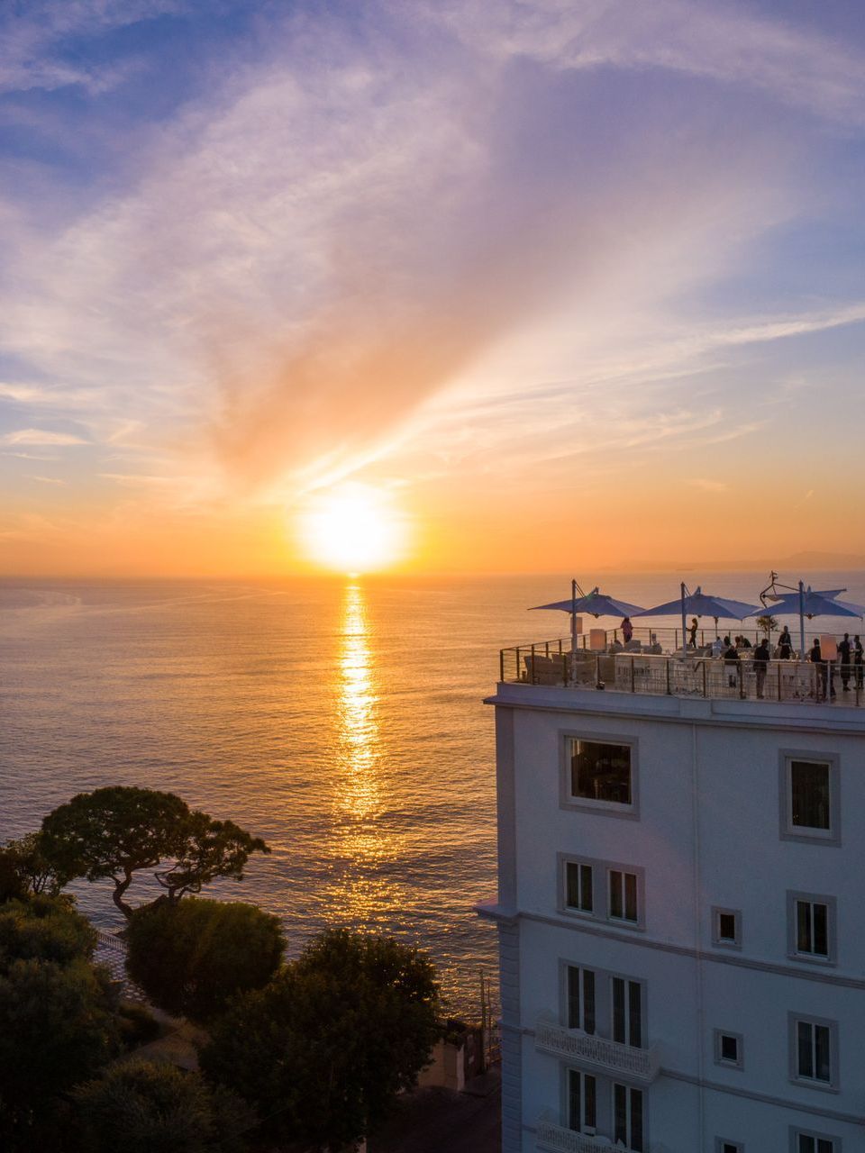 Il tramonto sull'oceano illumina un edificio costiero con una terrazza sul tetto affollata e ombrelloni blu.