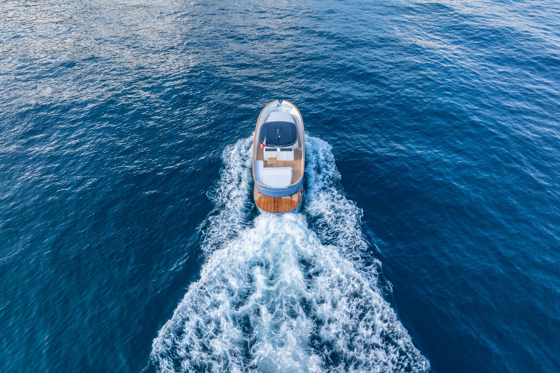 Aerial view of a small yacht cruising on deep blue ocean water, creating a white wake behind it.