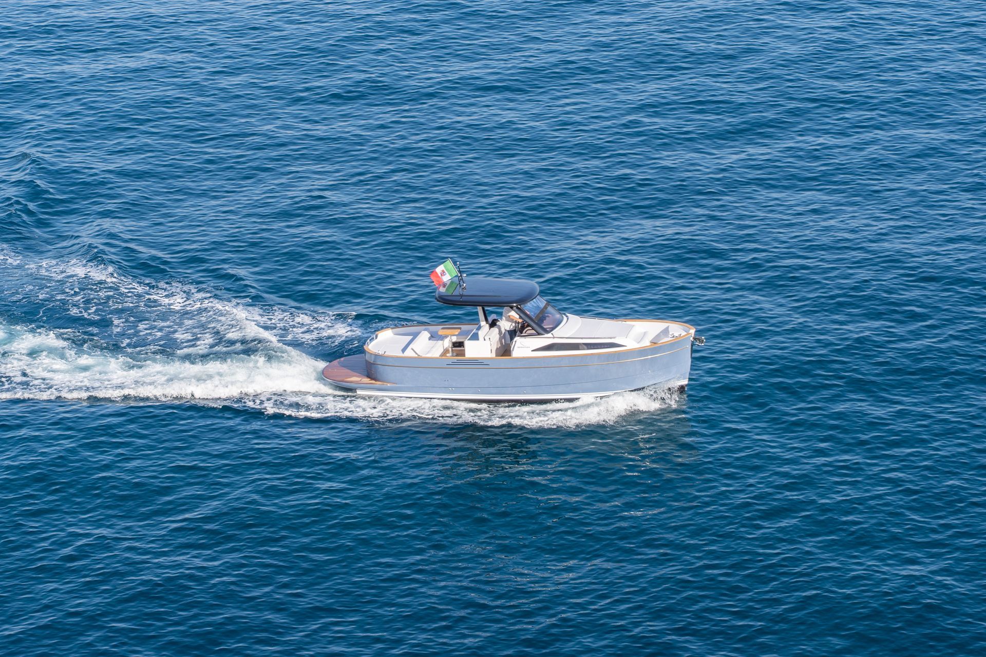 A small white motorboat with a blue canopy and an Italian flag cruises through deep blue water, leaving a wake.