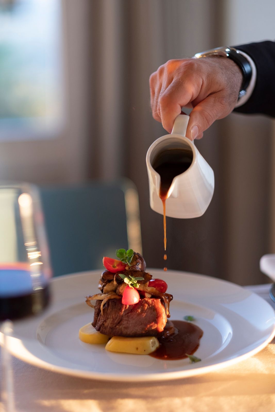 A hand pours brown sauce from a white creamer onto a small, stacked meat dish on a white plate.