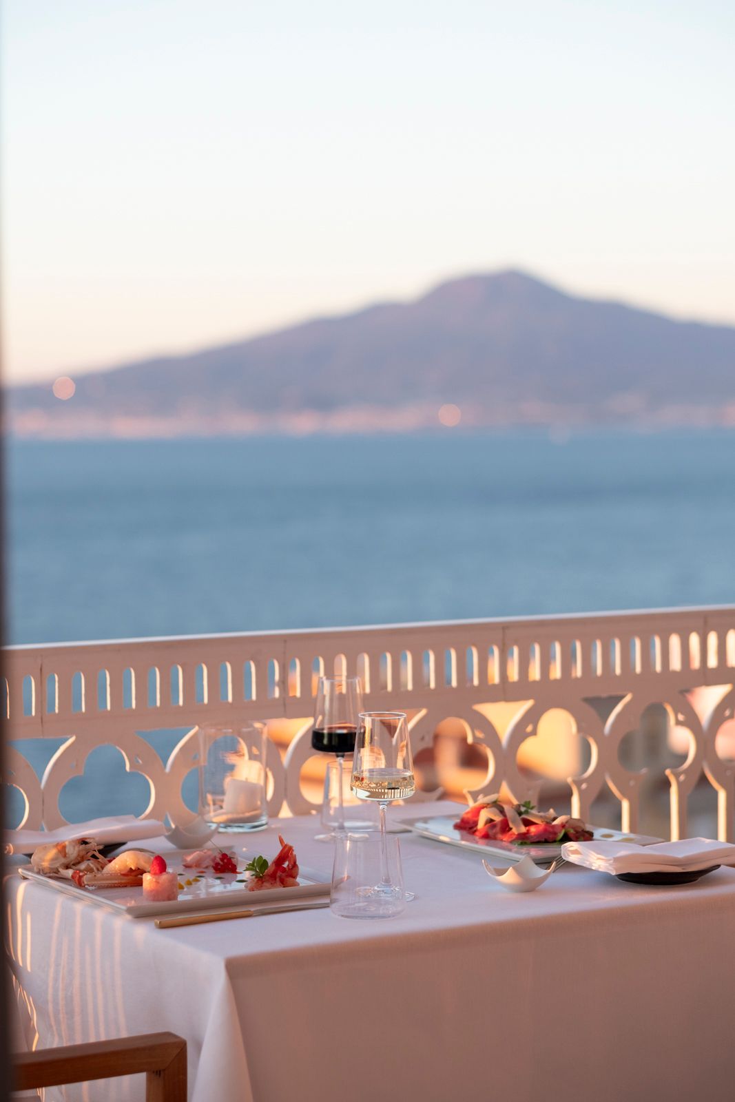 Un tavolo da pranzo apparecchiato per due su un balcone con vista sul mare e su un'imponente montagna in lontananza al tramonto.