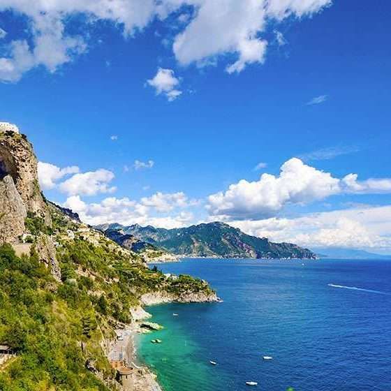 Scogliere scoscese ricoperte di vegetazione lussureggiante digradano verso un mare turchese e blu intenso, sotto un cielo limpido e soleggiato, punteggiato da soffici nuvole.