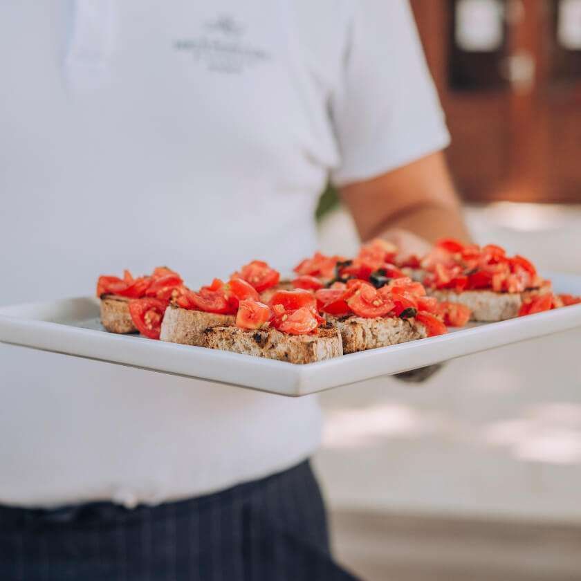 A waiter in a white uniform holding a rectangular platter of bruschetta topped with diced tomatoes.