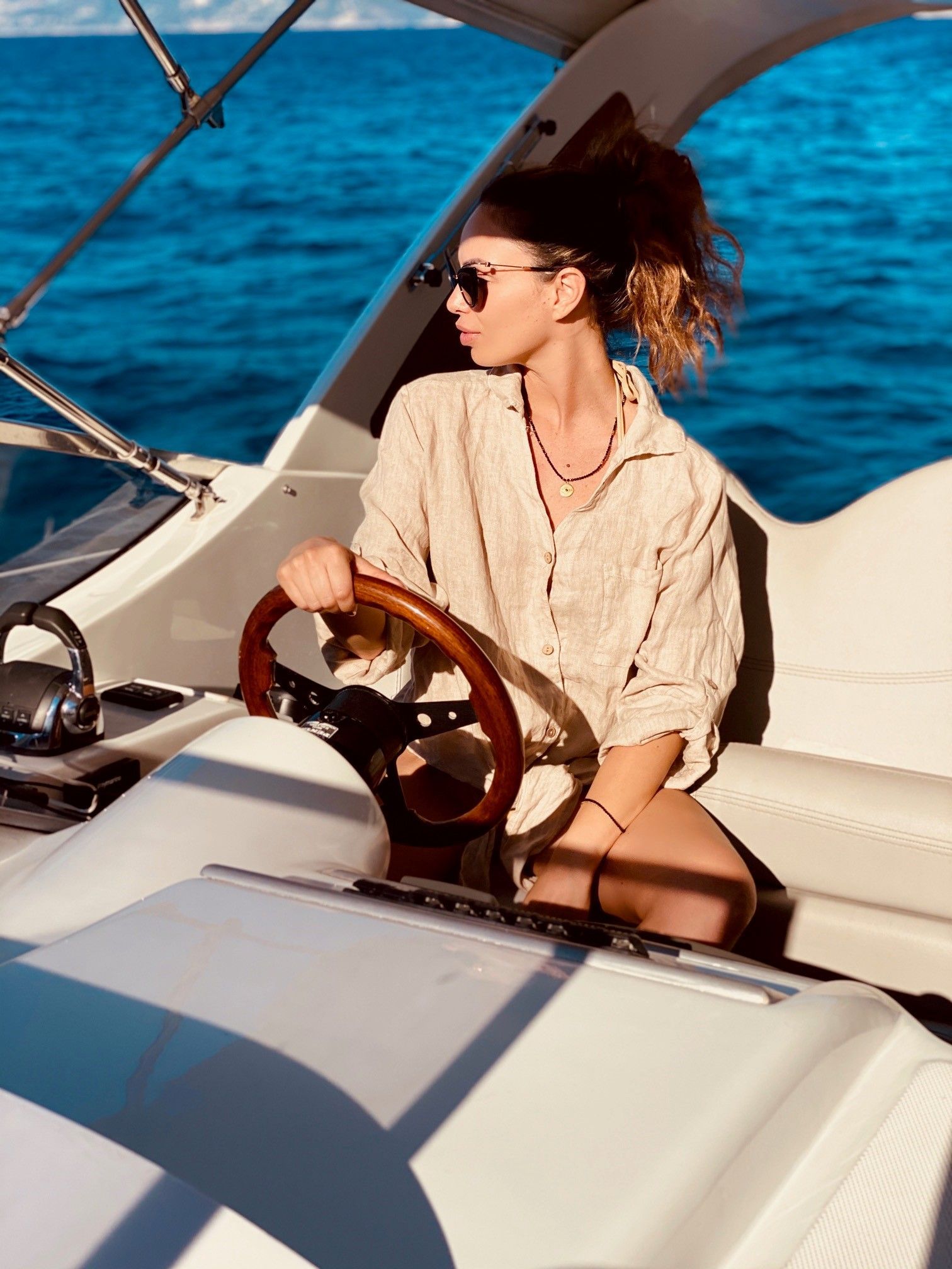 Una persona con una camicia di lino beige e occhiali da sole guida una barca su un mare blu brillante.