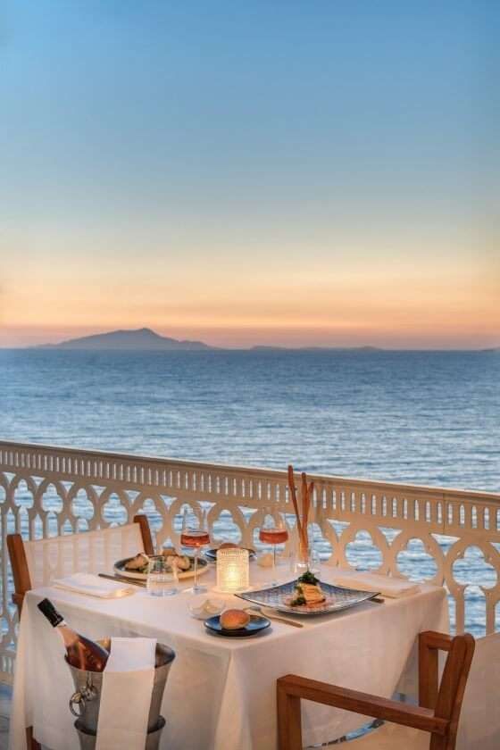 A romantic dinner table for two set on a balcony overlooking the ocean at sunset with a distant island on the horizon.