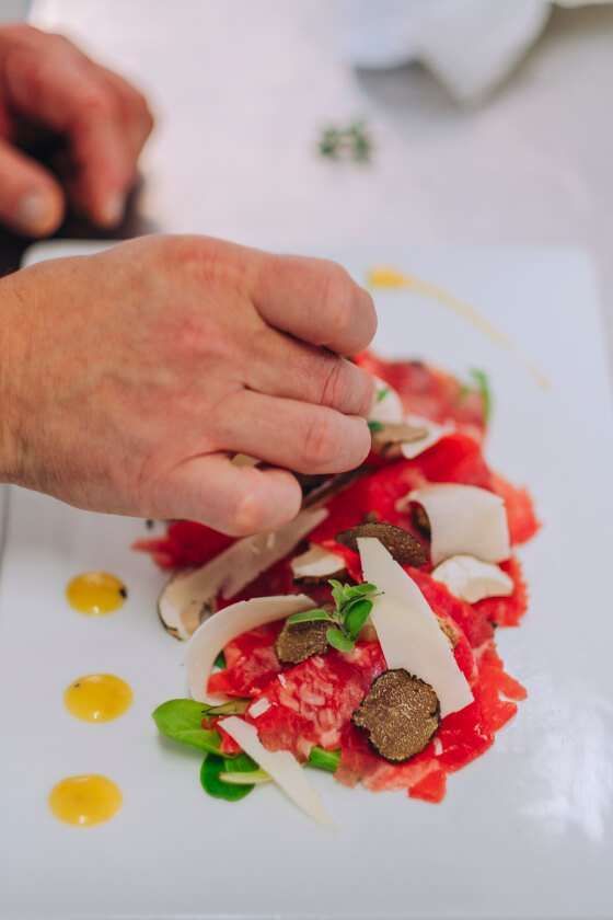 A close-up of a chef garnishing a plate of beef carpaccio topped with shaved cheese, fresh herbs, and truffle slices.