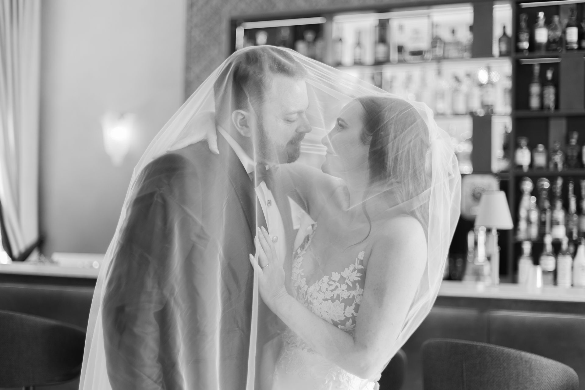 A bride and groom share a tender moment under a shared wedding veil inside a bar.