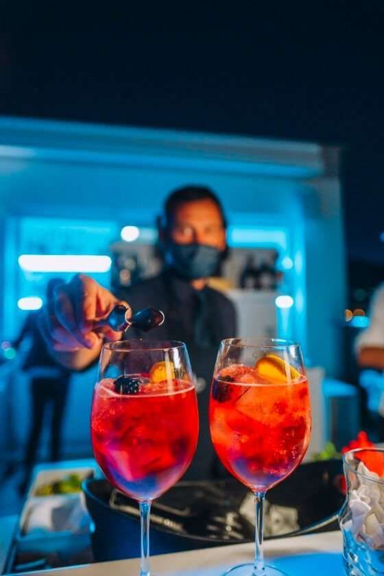 A person wearing a face mask places a garnish into one of two vibrant red cocktails at a bar with blue ambient lighting.