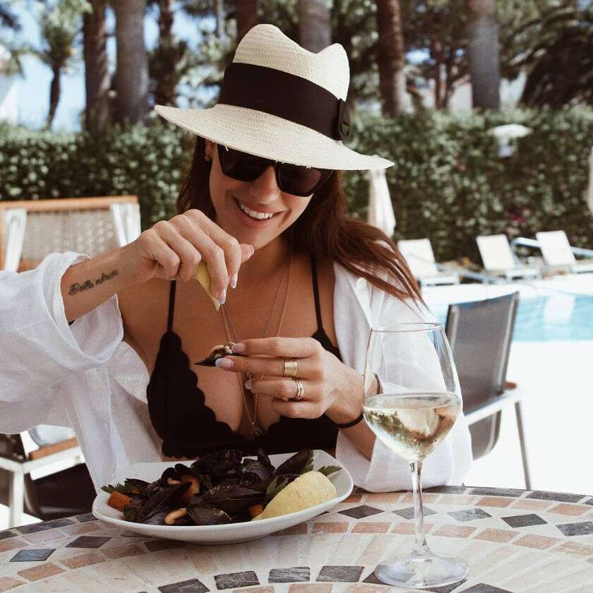 A person wearing a hat and sunglasses squeezes a lemon over a plate of mussels while sitting poolside with wine.