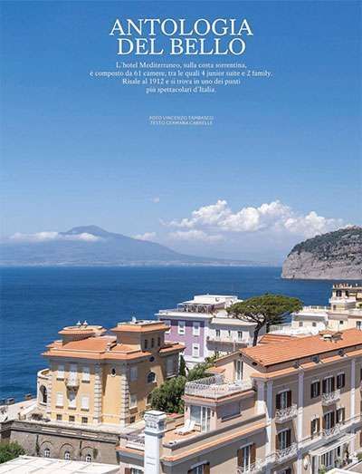 A sunny view of coastal buildings overlooking the sea, with Mount Vesuvius in the distance under a clear blue sky.