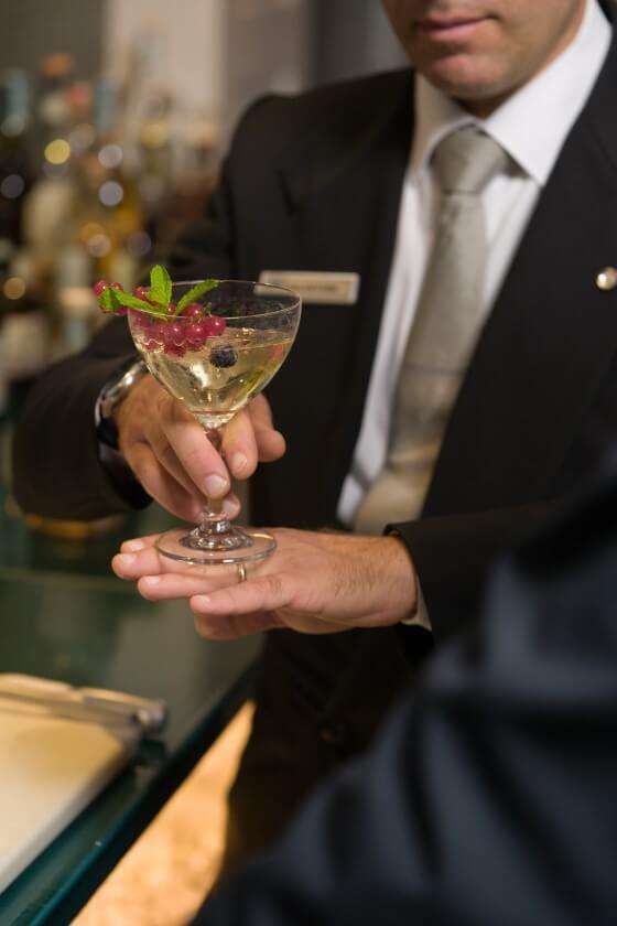 A server in a suit holding a stemmed cocktail glass garnished with berries and mint over a bar counter.
