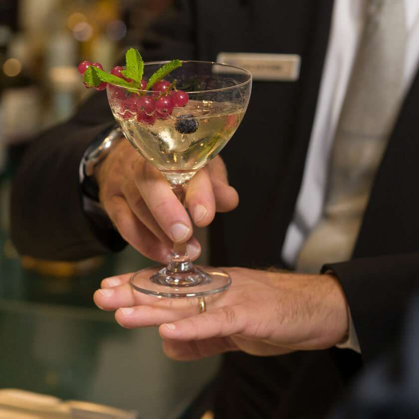A person in a suit serves a stemmed cocktail glass garnished with red currants and mint.