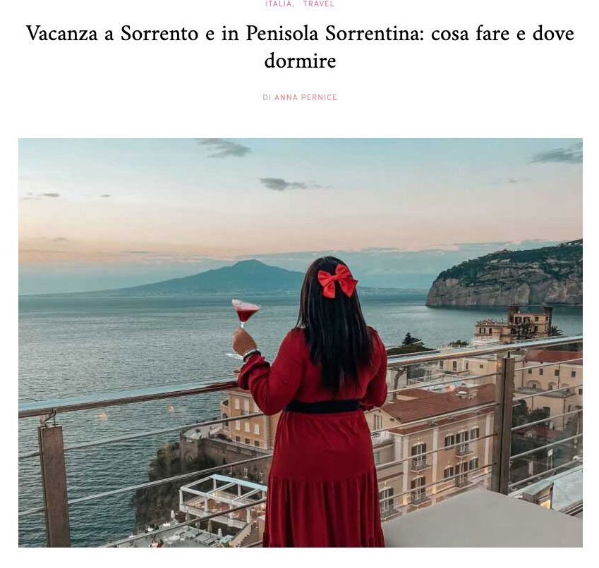 A person in a red dress with a red hair bow holds a cocktail overlooking a coastal town and sea at sunset.