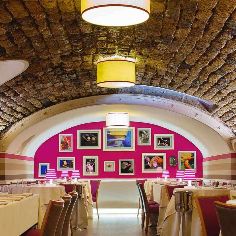 A dining room featuring a brick arched ceiling, tables with white cloths, and a bright pink wall decorated with photos.
