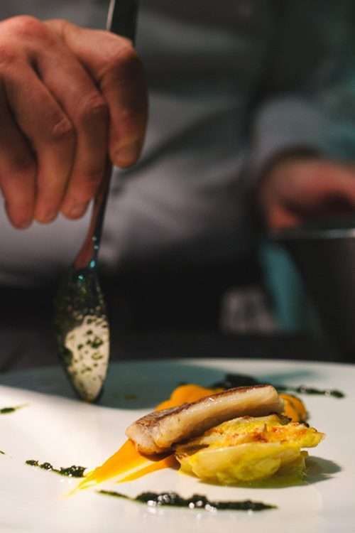A chef's hand uses a spoon to plate a piece of seared fish over vegetables and colorful sauces on a white plate.