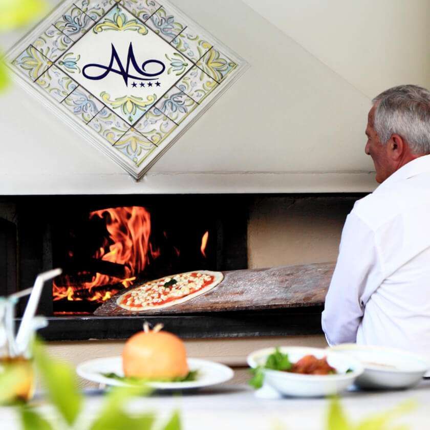 A chef uses a wooden peel to place a pizza into a wood-fired oven. Plated dishes sit on a counter in the foreground.