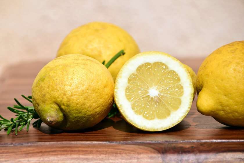 Three whole lemons and one halved lemon displayed on a wooden surface with a sprig of fresh rosemary.