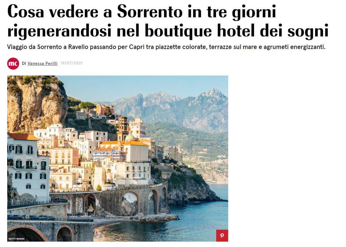 A sunlit coastal Italian town with colorful buildings perched on cliffs above the sea against a mountain backdrop.