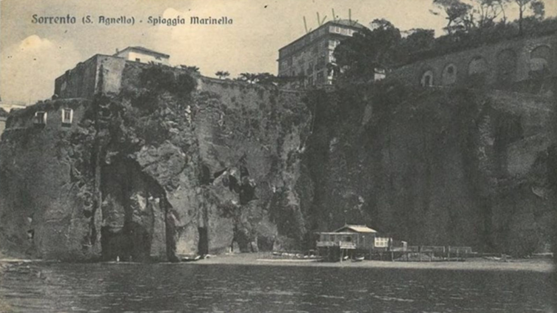 Vintage postcard showing a small beach at the base of tall, rugged cliffs in Sorrento, Italy, with a wooden pier.