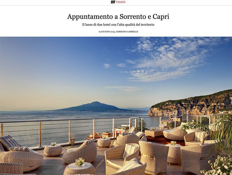 Wicker patio furniture overlooking a calm sea with a mountain silhouette on the horizon under a sunny sky.