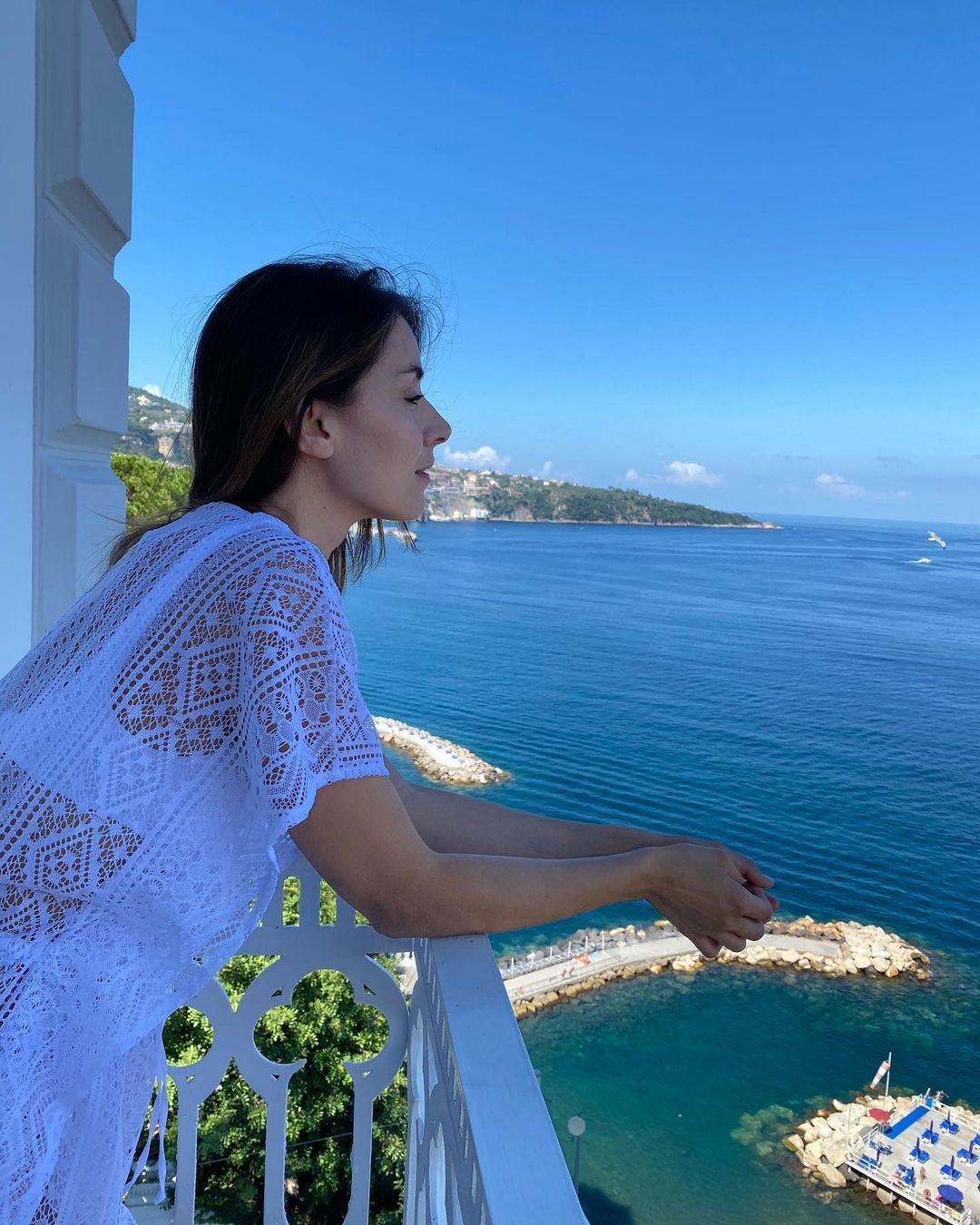 Una persona con indosso un copricostume di pizzo bianco si appoggia alla ringhiera di un balcone, guardando l'oceano, una diga frangiflutti e il cielo azzurro.