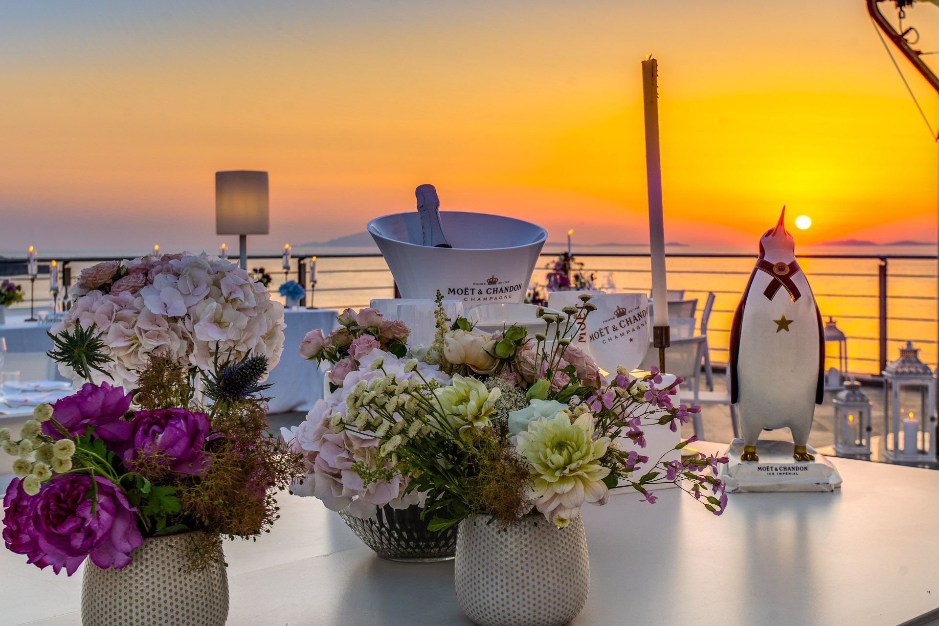 A sunset view over the ocean with decorative floral arrangements, a wine cooler, and a penguin statue on a table.