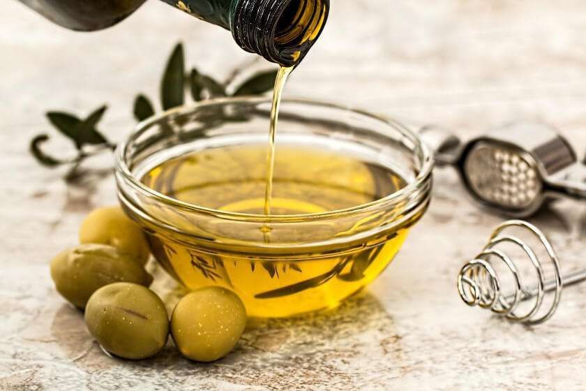 Olive oil pours from a bottle into a clear glass bowl, surrounded by whole green olives and kitchen tools on a marble base.
